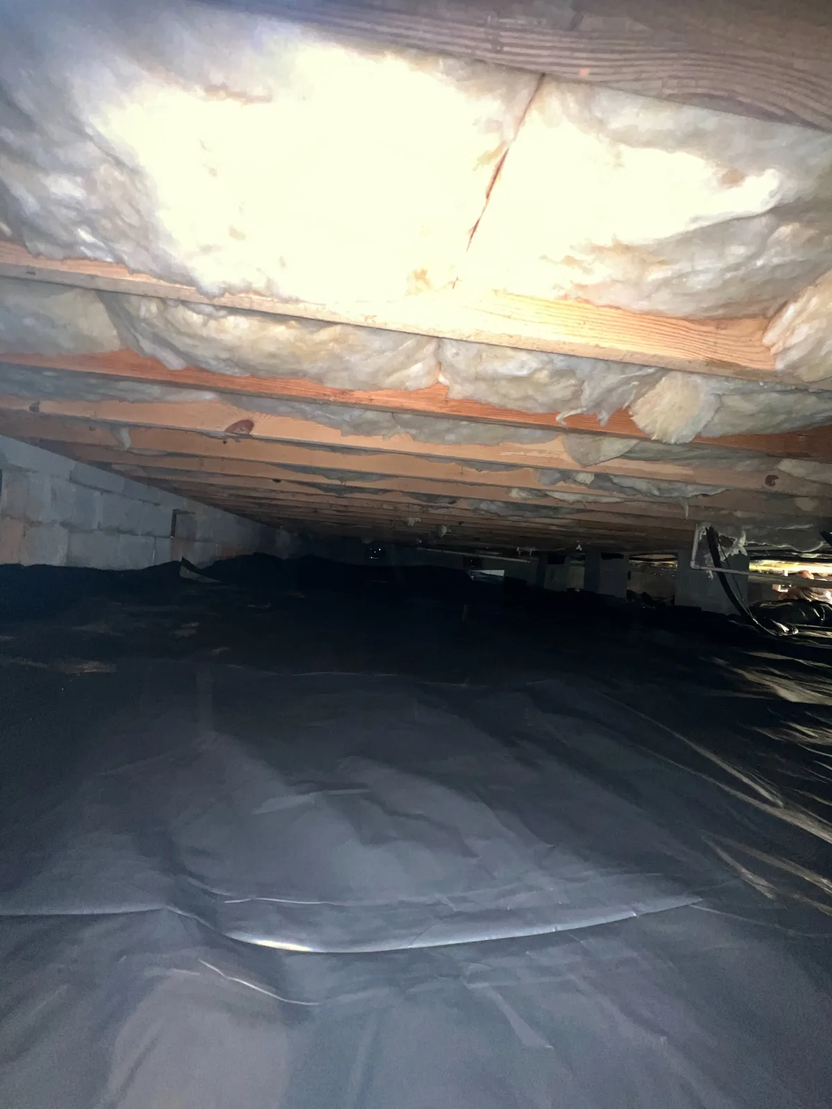 Same crawlspace area with a black vapor barrier installed and drain piping in place
